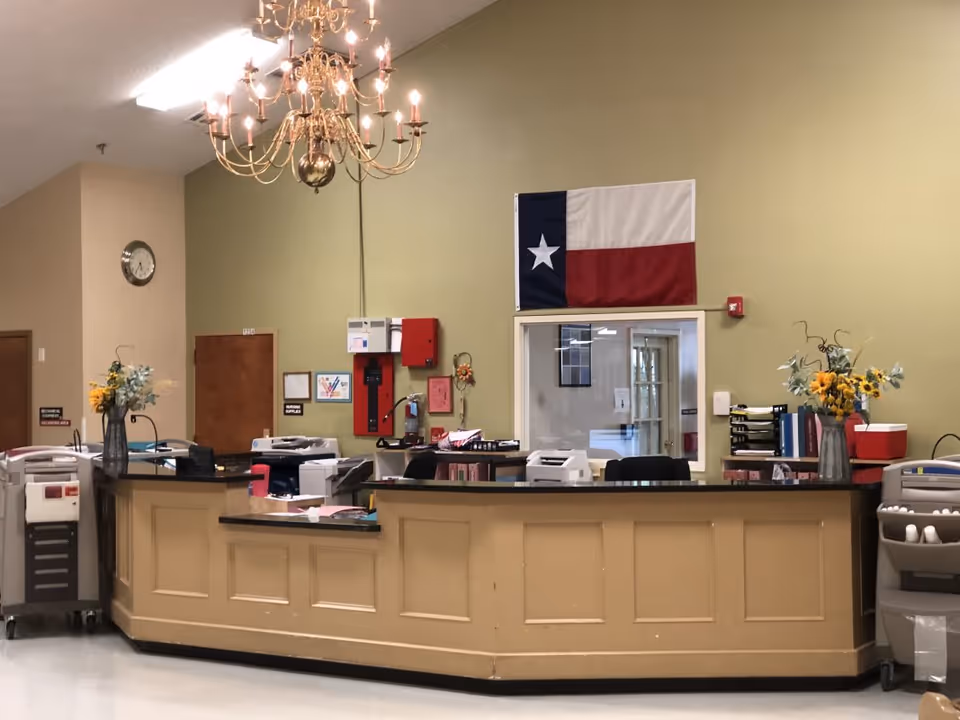 Reception desk area inside a facility with a beige counter, two vases with yellow flowers, office equipment, and a Texas state flag hanging on the wall behind the desk. A chandelier is hanging from the ceiling, and there are doors and a clock on the wall in the background.