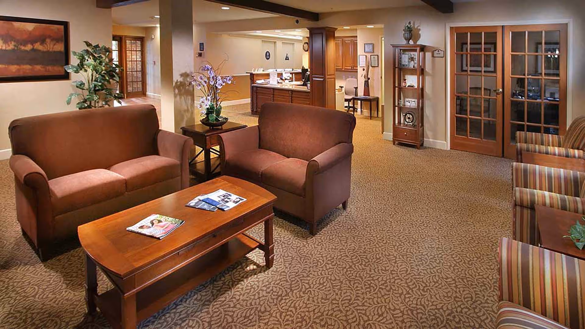 A cozy senior living facility lounge area with two brown upholstered loveseats facing a wooden coffee table with magazines on it. There are striped armchairs on the right side, a wooden display cabinet against the wall, and a reception desk in the background. The room has beige walls, patterned carpet, and framed artwork and plants for decoration.