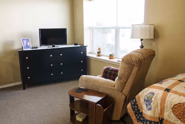 A cozy room with a beige recliner chair next to a wooden side table holding a TV remote, a black dresser with a flat-screen TV and a framed photo on top, a window with blinds and small potted plants on the windowsill, and part of a bed with a colorful quilt.