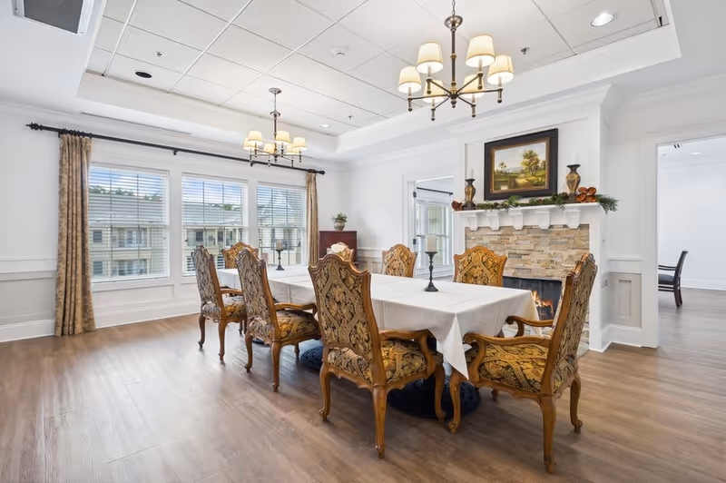 A dining room with a long table covered with a white tablecloth and surrounded by eight ornate wooden chairs with patterned upholstery. The room features a stone fireplace with a painting and decorative items on the mantel. Two chandeliers hang from the ceiling, and large windows with curtains allow natural light to fill the space.