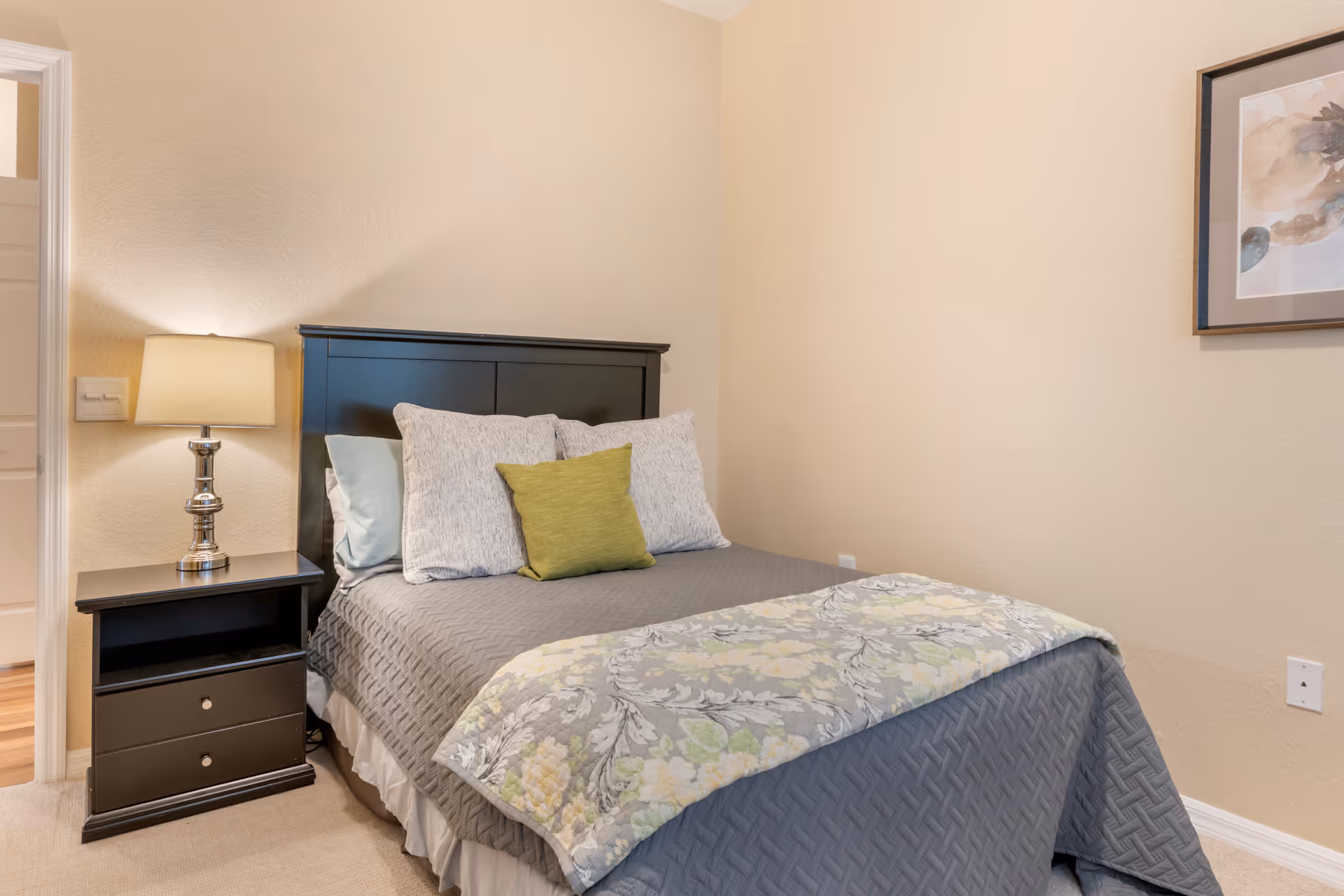 Small bedroom with a made bed featuring grey bedding and decorative pillows, a black nightstand with a lamp, and framed wall art.