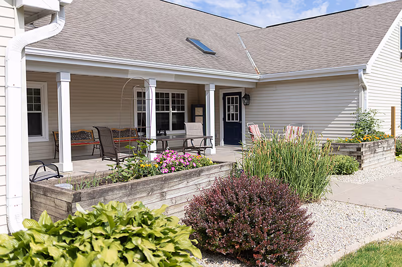 Covered front patio of a senior living facility with seating, raised wooden planters, and landscaping.