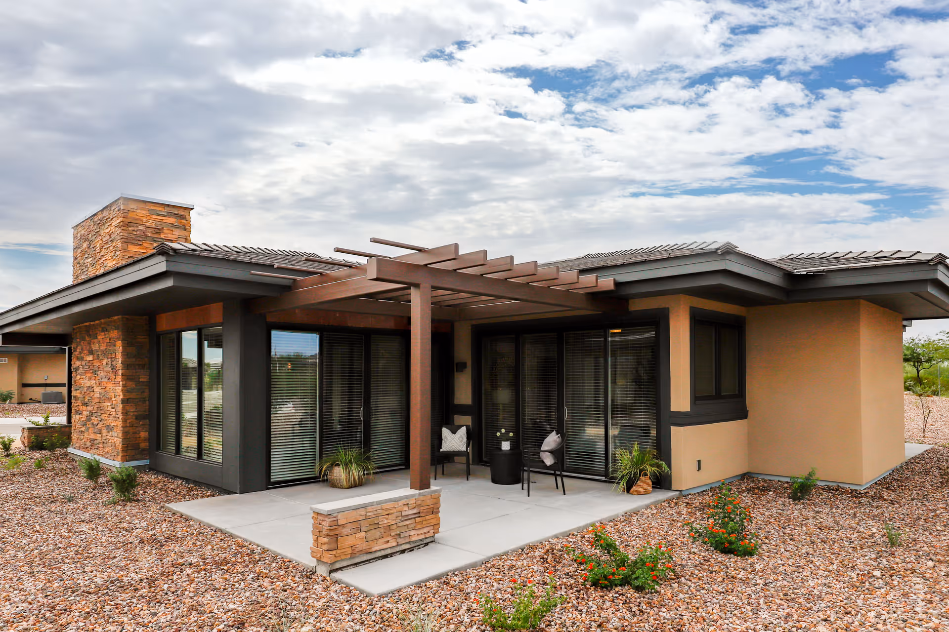 Exterior view of a modern single-story building with a stone chimney and large windows. The building features a covered patio area with a wooden pergola, two chairs, a small table, and some potted plants. The surrounding landscape is covered with gravel and small shrubs under a partly cloudy sky.