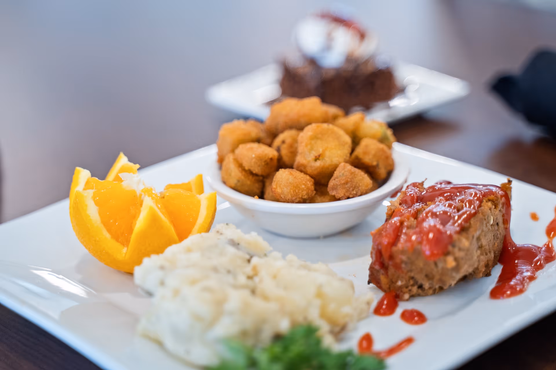A close-up of a white plate with a serving of mashed potatoes, a slice of meatloaf topped with ketchup, a bowl of fried okra, and orange slices. In the background, there is a dessert plate with a brownie and whipped cream.