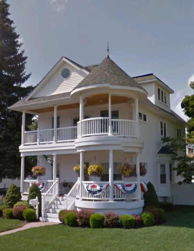A large white two-story house with a wraparound porch and a rounded turret-like balcony on the second floor. The porch is decorated with hanging flower baskets and patriotic red, white, and blue bunting. The house is surrounded by neatly trimmed bushes and a green lawn, with a large tree on the left side.