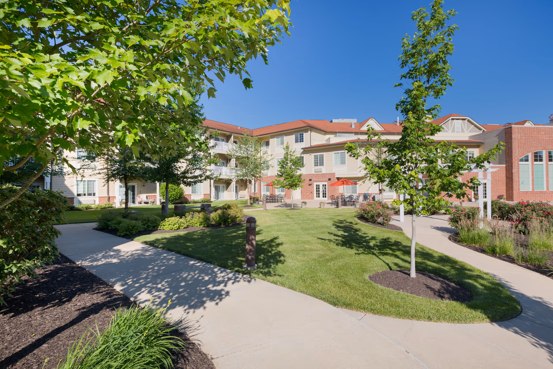 Outdoor courtyard area of Crestview Senior Living with well-maintained green lawns, young trees, shrubs, and a paved walkway. The building has a beige and red brick exterior with multiple windows and balconies. There are outdoor seating areas with tables and umbrellas under a clear blue sky.
