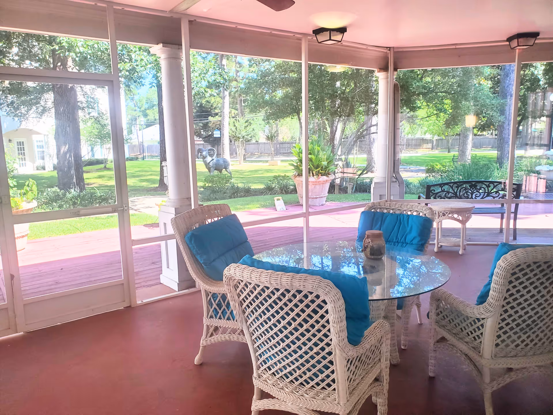A bright enclosed patio area with a round glass table surrounded by four white wicker chairs with blue cushions. Large windows provide a view of a green garden with trees, plants, and a decorative ram statue. The floor is reddish, and there are ceiling lights and a fan above.