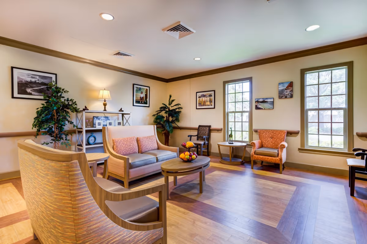 A bright and spacious living room in Magnolia Estates Rehab & Retirement featuring wooden flooring, a beige sofa with two pink cushions, two armchairs, a coffee table with a bowl of fruit, a side table with a small decorative item, framed pictures on the walls, two large windows letting in natural light, and potted plants adding greenery to the space.