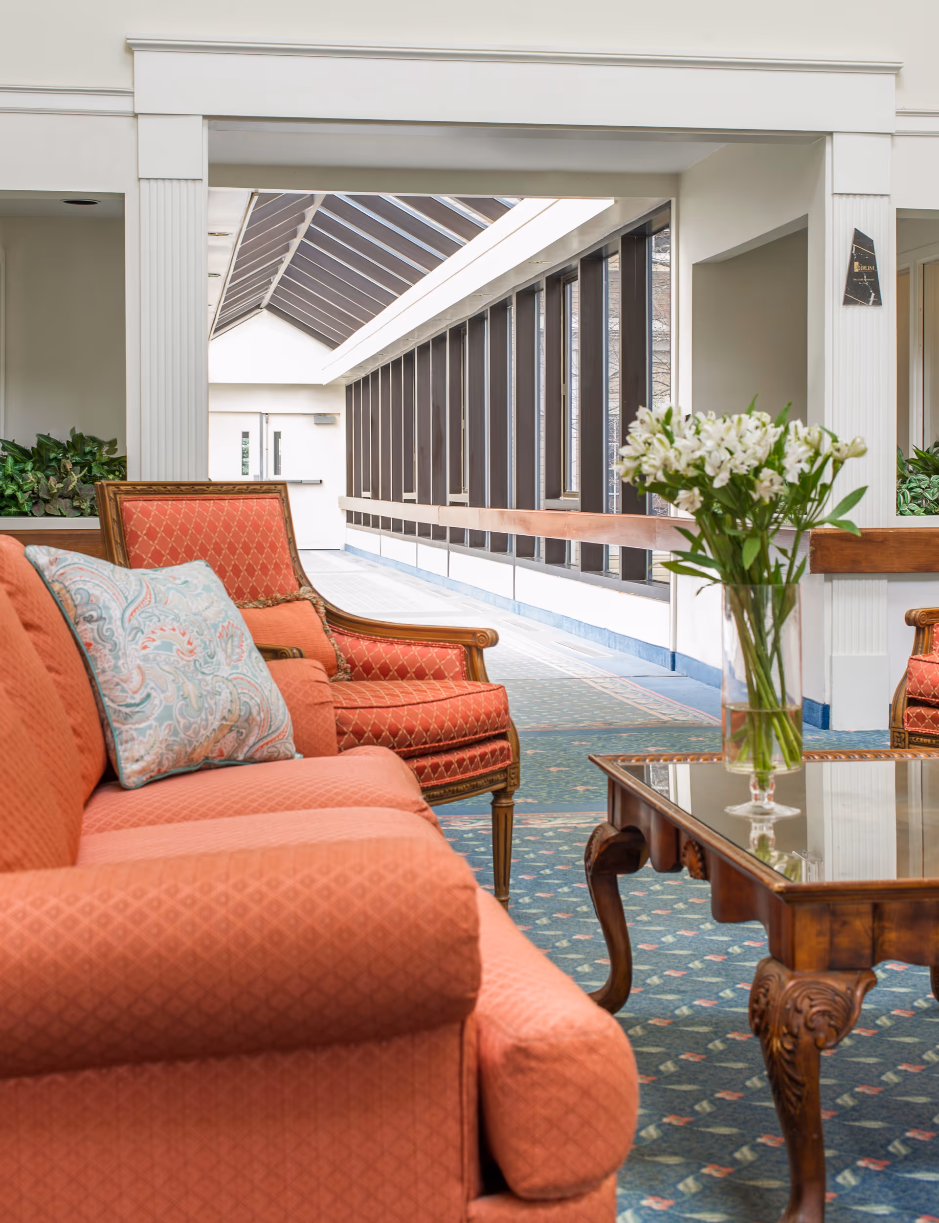 A cozy indoor seating area with coral-colored upholstered armchairs and a matching sofa, a wooden coffee table with a glass top holding a vase of white flowers, and a long corridor with large windows and a skylight ceiling in the background.