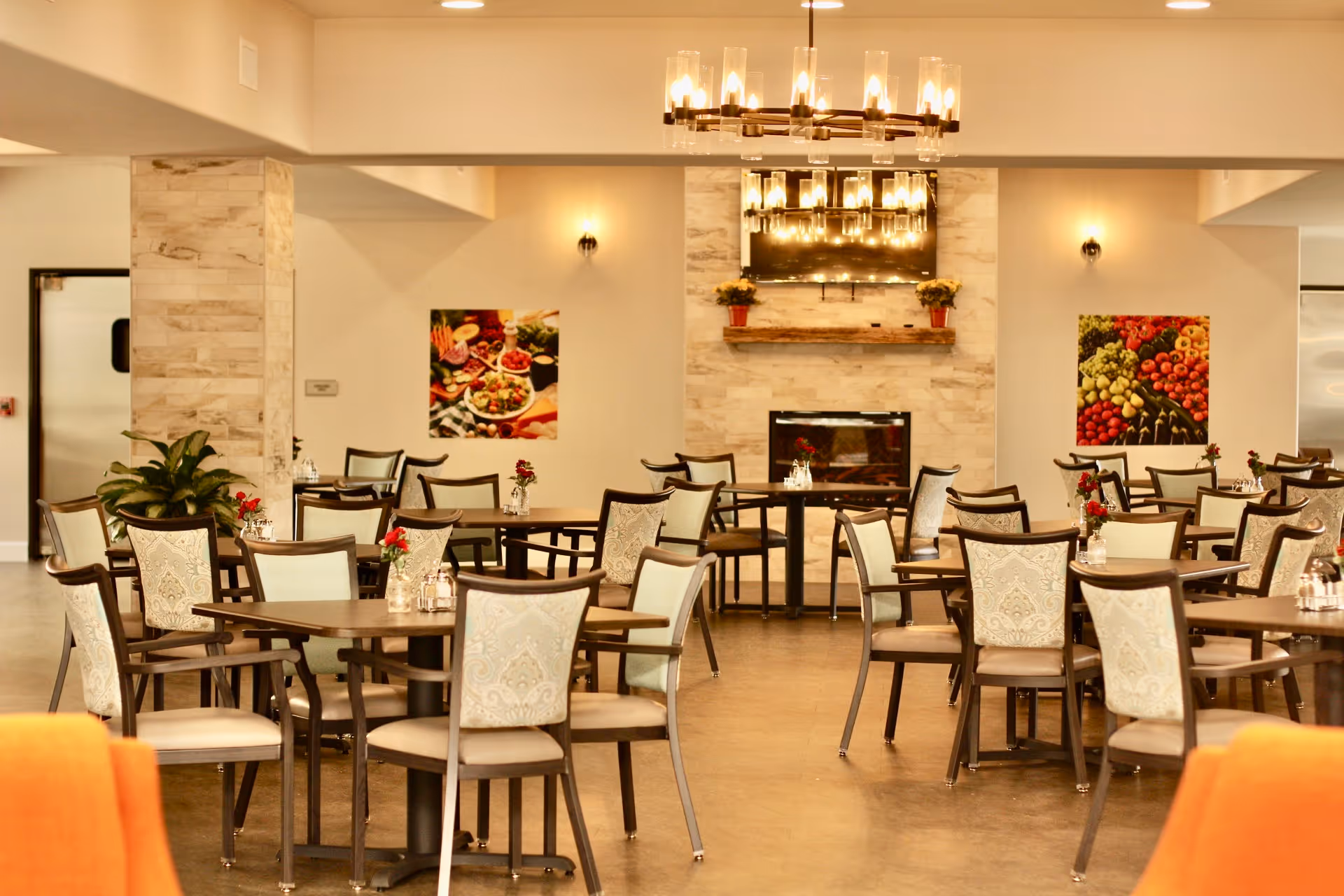 A spacious dining room with multiple wooden tables and cushioned chairs arranged neatly. The room features a modern chandelier, a fireplace with a mounted TV above it, and colorful food-themed artwork on the walls. Small vases with red flowers are placed on each table, and there are plants near the walls.