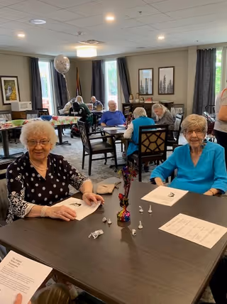 A group of elderly people sitting at tables in a well-lit common room. Two elderly women are seated at the front table, smiling and holding papers. Other seniors are seated at tables in the background, engaged in activities. The room has large windows with curtains, framed artwork on the walls, and a festive balloon in the background.