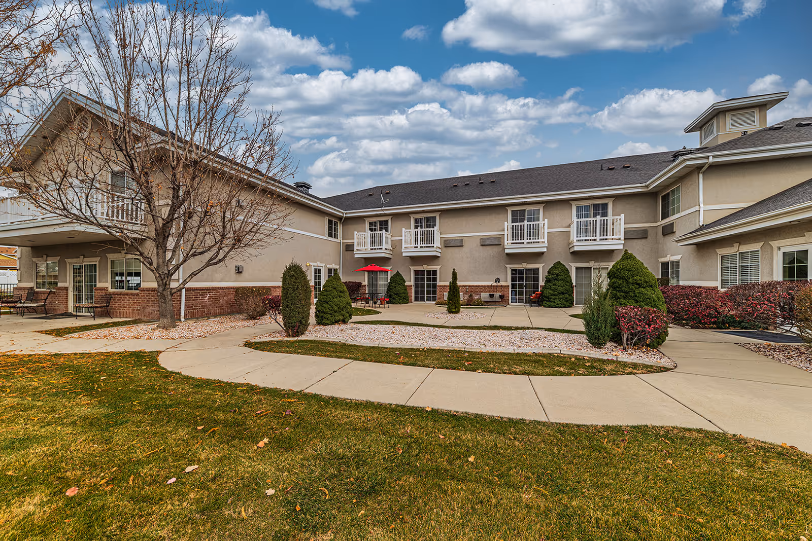 Exterior view of a senior living facility building with beige walls and a dark roof under a partly cloudy sky. The building has multiple windows and small balconies. There is a paved walkway surrounded by green grass, shrubs, and small trees, with a red umbrella shading a patio table and chairs near the center.