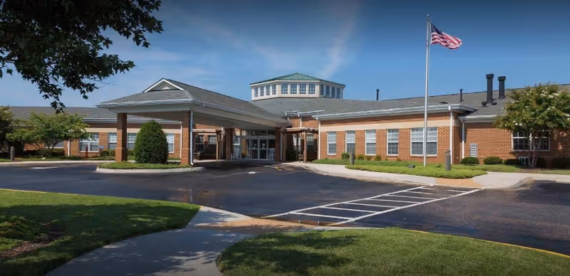 Brick single-story assisted living building with a covered entrance, American flag, and circular driveway under a blue sky.