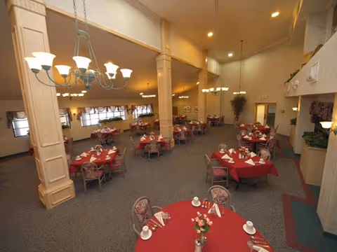 Spacious dining room with multiple round tables covered in red tablecloths, each set with white napkins, cups, and small flower arrangements. The room features high ceilings, chandeliers, large windows with curtains, and beige walls with decorative columns.