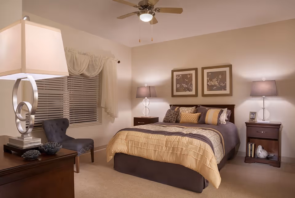 A cozy bedroom with a large bed featuring a yellow and brown comforter and multiple pillows. There are two matching nightstands on either side of the bed, each with a lamp. Above the bed are two framed floral artworks. A window with blinds and sheer curtains is on the left wall, next to a dark upholstered chair. A modern table lamp sits on a dresser in the foreground.