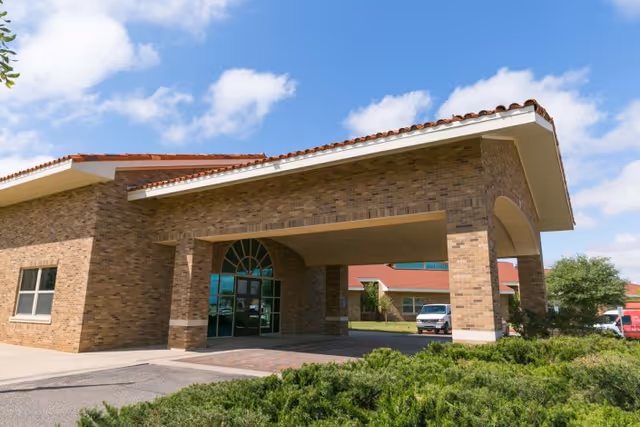Exterior view of The Garrison Geriatric Education and Care Center showing a brick building with a covered entrance supported by pillars, a tiled roof, and a clear blue sky with some clouds. There are shrubs in the foreground and a few vehicles parked near the building.