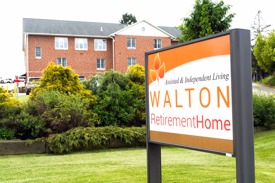 A large sign for Walton Retirement Home with the text 'Assisted & Independent Living WALTON Retirement Home' in front of a landscaped area with bushes and a brick building in the background.