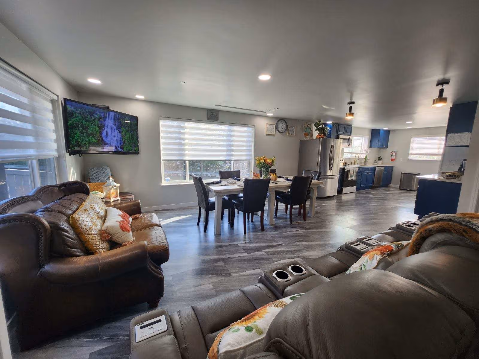 A spacious and modern open-concept living area featuring a brown leather sofa with decorative pillows, a reclining leather chair with cup holders, a dining table with six chairs, and a kitchen with blue cabinets, stainless steel appliances, and large windows with blinds allowing natural light to fill the room.