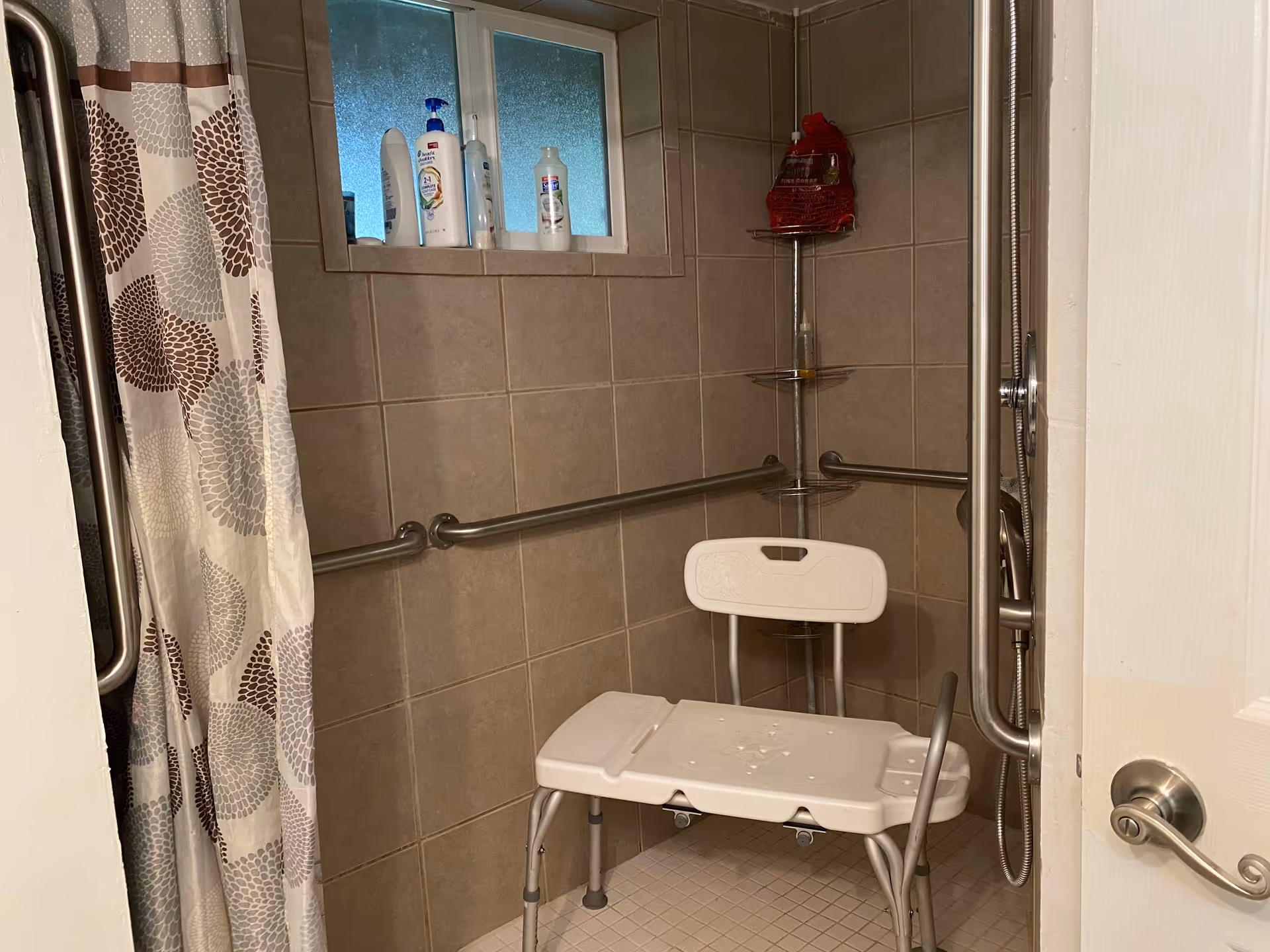 Shower area with beige tiled walls and floor, equipped with a white shower chair, metal grab bars on the walls, a shower curtain with a floral pattern in shades of brown and gray, and a small frosted window with various bottles on the sill.