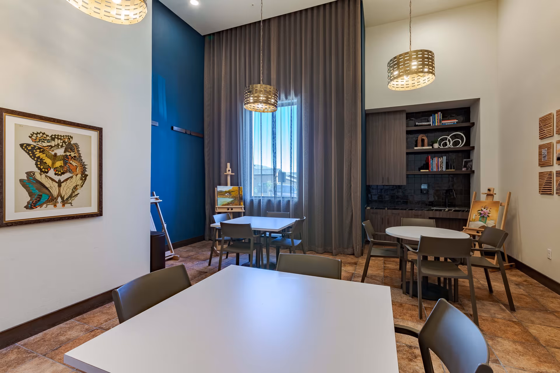 Bright communal room with several tables and chairs, pendant lights, a kitchenette with shelves, and artwork including easels and a framed butterfly print.