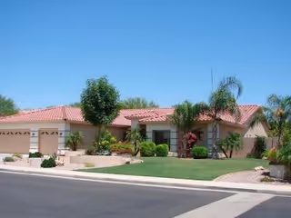 Single-story residential building with a red tile roof, beige exterior walls, and a well-maintained front yard featuring green grass, palm trees, and other plants under a clear blue sky.