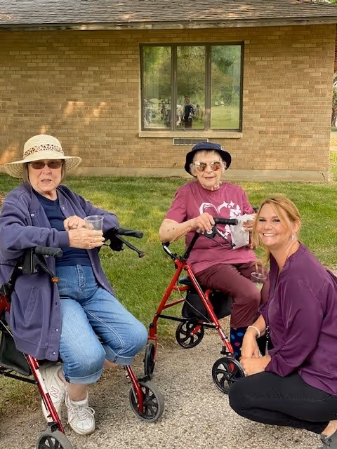 Two elderly women sitting outdoors on walkers, one wearing a sun hat and sunglasses, the other wearing a bucket hat and sunglasses, both holding drinks. A younger woman in a purple top is crouching beside them, smiling. They are outside near a brick building with a window reflecting greenery.