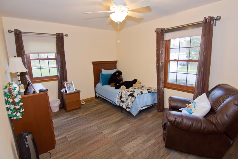 A cozy bedroom with a single bed covered in a blue blanket and a patterned throw. There are two stuffed animals on the bed. The room has two windows with brown curtains, a ceiling fan with light, a brown leather armchair with a stuffed toy, a wooden dresser with a lamp and framed photos, and a small wooden nightstand.
