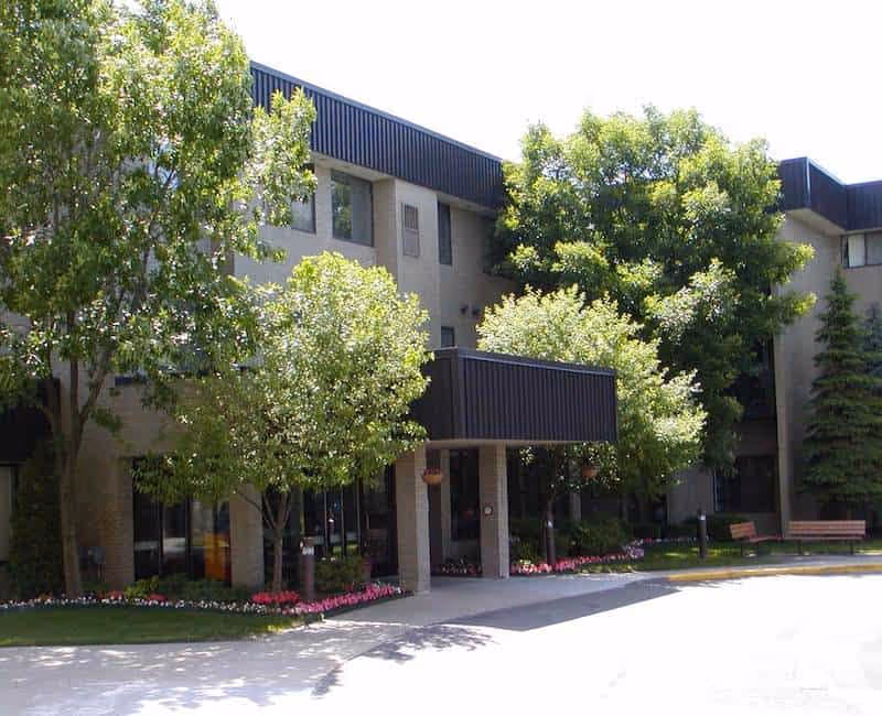 Exterior view of a multi-story senior living facility building with beige walls and dark trim. The entrance is covered by a black awning, surrounded by green trees and landscaped flower beds with pink flowers. There are benches on the lawn near the entrance.