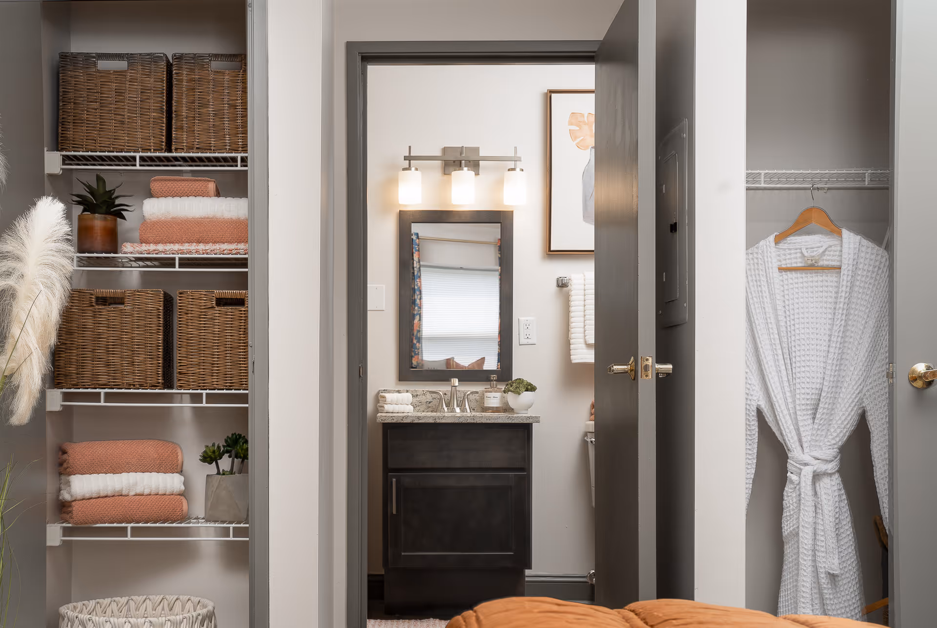 View of a bathroom vanity with a mirror and three-light fixture above it, seen through an open doorway. To the left of the doorway is a shelving unit with wicker baskets, folded towels, and a small plant. To the right is a closet with a white robe hanging on a wooden hanger.
