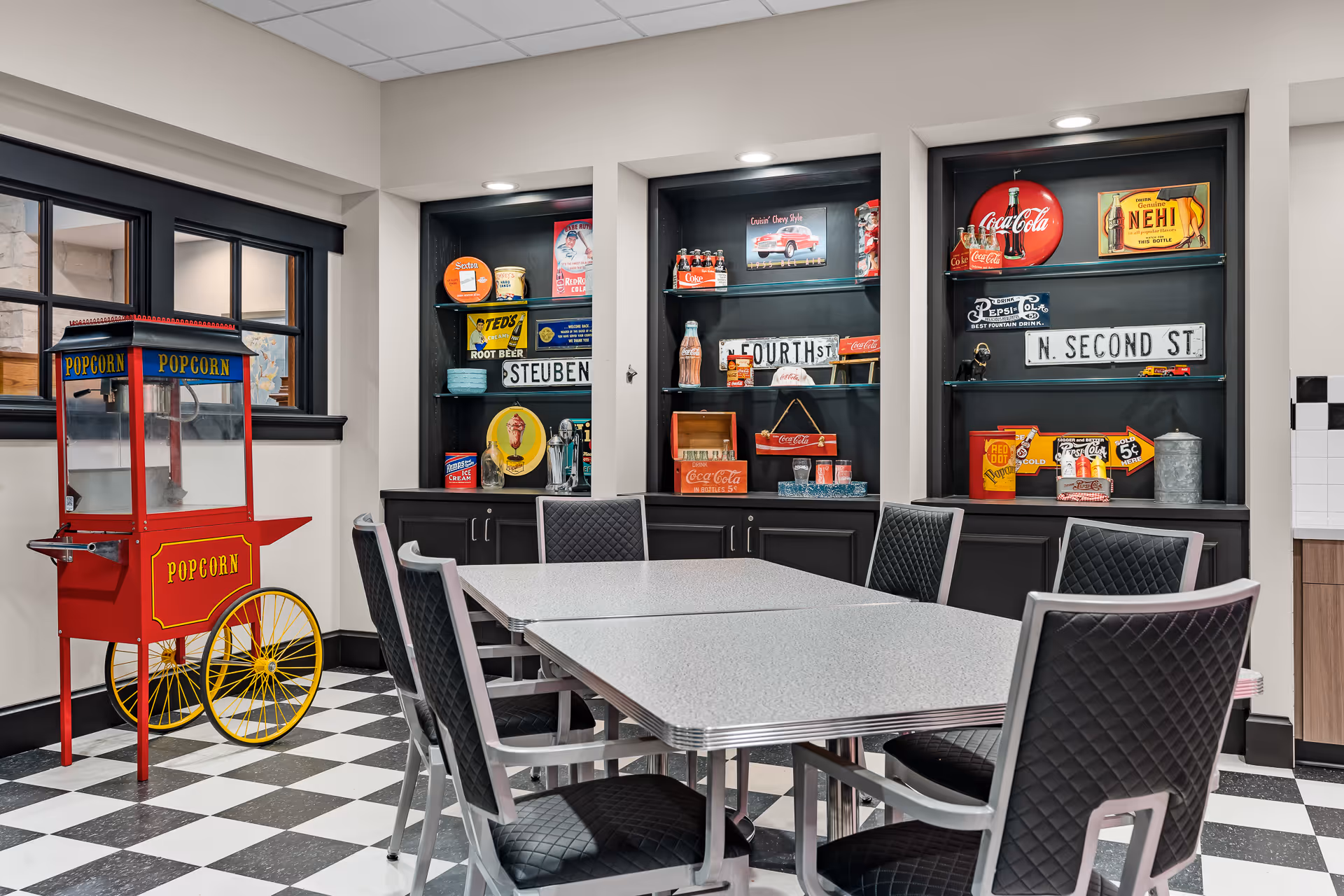 A retro-themed dining area with a black and white checkered floor, a red vintage popcorn cart, and a table surrounded by six black cushioned chairs. Behind the table are built-in black shelves displaying vintage soda and snack memorabilia, including Coca-Cola and Pepsi signs, old street signs, and various nostalgic items.