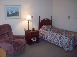 A small bedroom with a single bed covered in a floral bedspread, a wooden nightstand with a lamp and a phone, and a plush armchair. A framed picture of a beach scene hangs on the wall above the armchair.