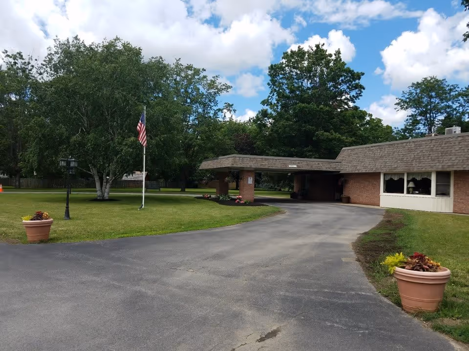Exterior view of Absolut Care of Aurora Park facility showing a driveway leading to a covered entrance. There are green lawns with trees, an American flag on a flagpole, two large flower pots with plants, and a partly cloudy sky.