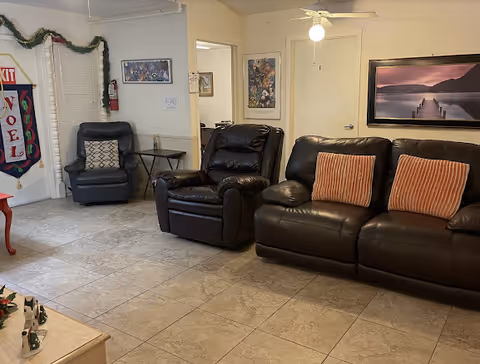 A cozy living room area with a dark leather sofa adorned with two orange striped pillows and a matching leather recliner chair. Another recliner chair with a patterned cushion is placed near a window decorated with a holiday garland and a 'NOEL' banner. The room has tiled flooring, a ceiling fan with a light, framed artwork on the walls, and a small side table with a bottle on it.