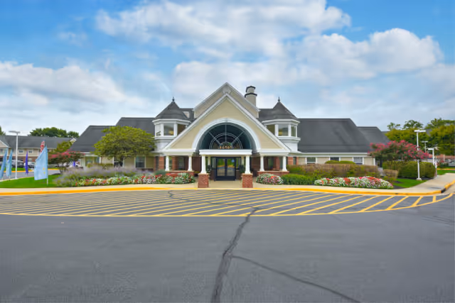 Front exterior view of a senior living facility named Cascades Village with a large entrance featuring an arched window, two small turret-like structures on the roof, well-maintained flower beds, and a spacious parking area with yellow striped markings.