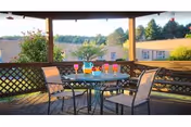 Outdoor patio area with a round glass table and four chairs. On the table are several colorful drinks and a pitcher. The patio has wooden railings with lattice panels and overlooks a building and greenery in the background under a clear sky.