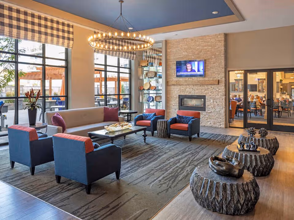 Bright contemporary lounge with sofas and armchairs around a coffee table, a stone fireplace with a wall-mounted TV, and large windows looking out to a patio.