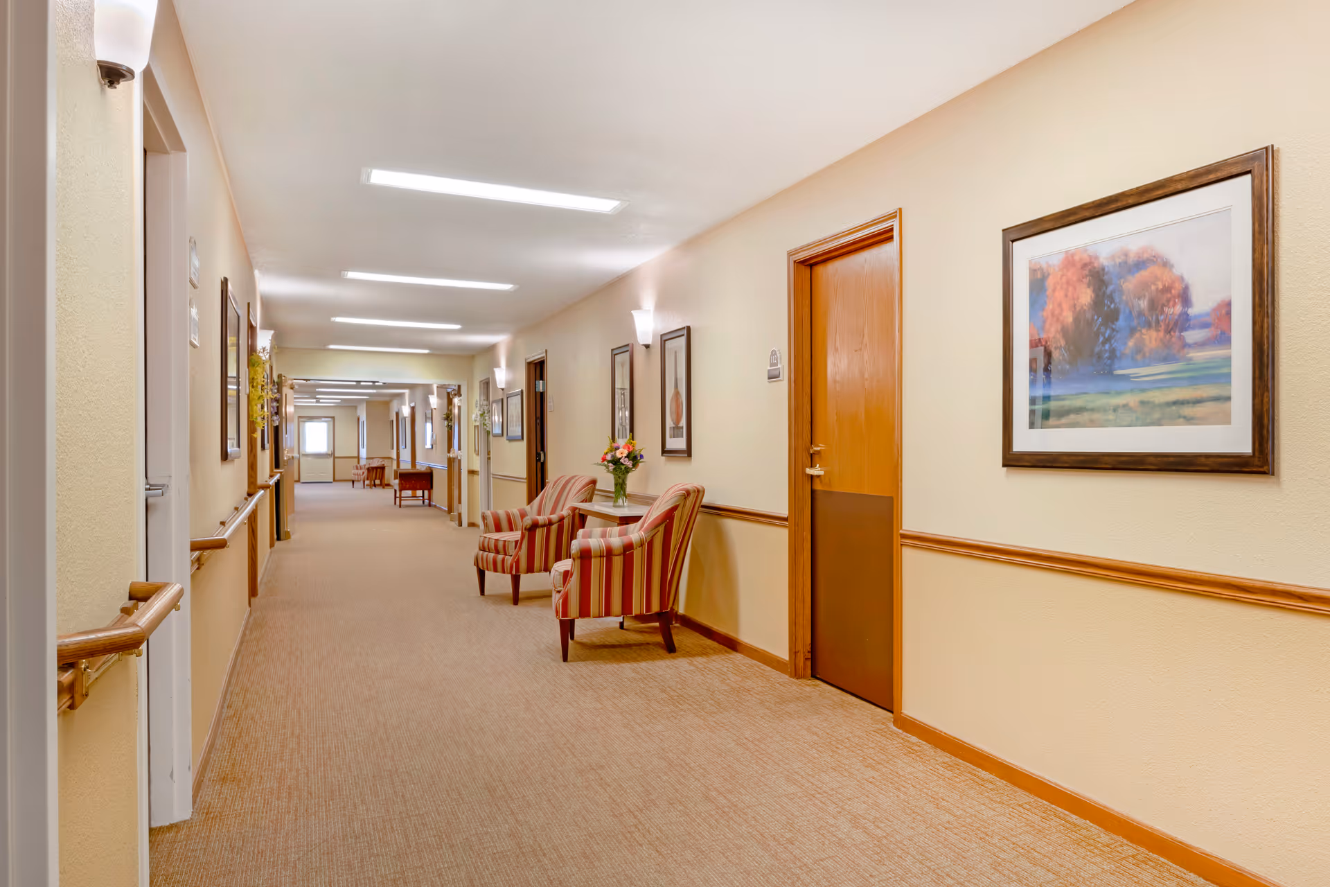 A well-lit hallway in a senior living facility with beige walls and carpeted floor. The hallway features wooden handrails on both sides, framed artwork on the walls, and several wooden doors. Two striped armchairs with a small table holding a vase of flowers are placed along the right side of the hallway. Wall-mounted light fixtures provide additional lighting.