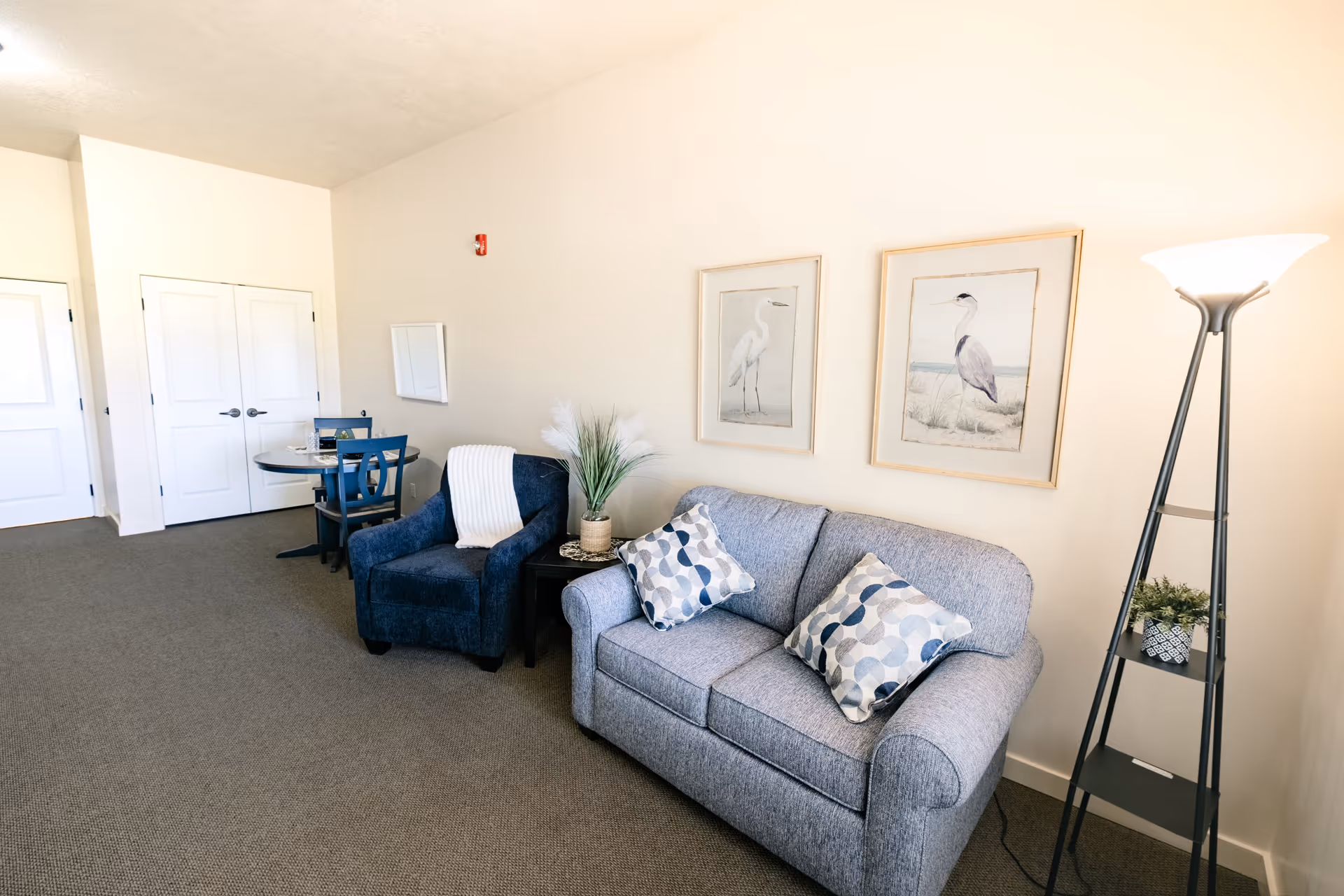 A cozy living room area with a light blue sofa adorned with two patterned pillows, a dark blue armchair with a white throw blanket, a small black side table with a vase of decorative grass, two framed bird prints on the wall, a tall floor lamp with a bright light, and a small round dining table with two chairs near white double doors.