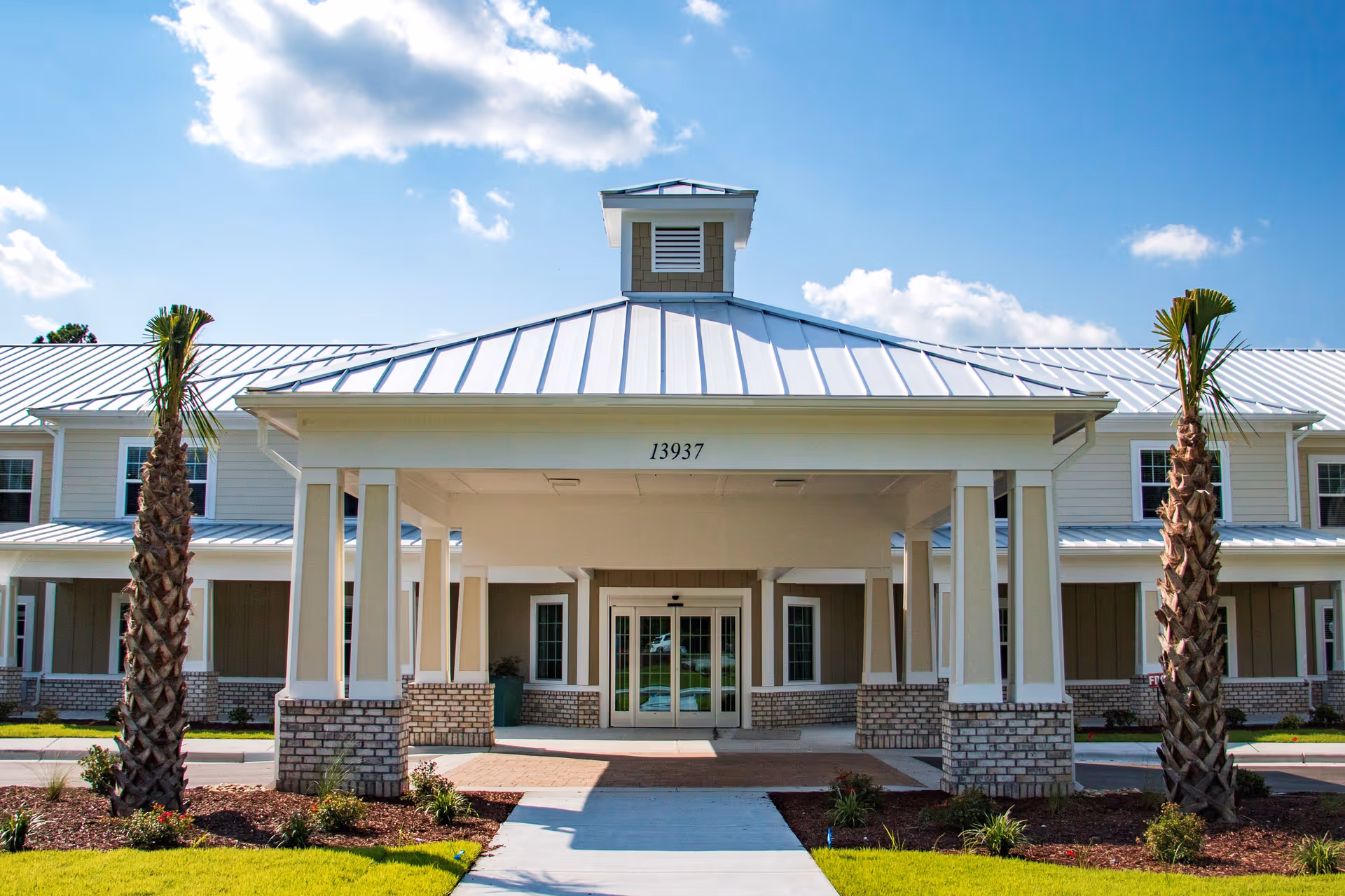 Front exterior view of Arbor Landing at Hampstead building with a covered entrance supported by columns, two palm trees, and a clear blue sky with some clouds.