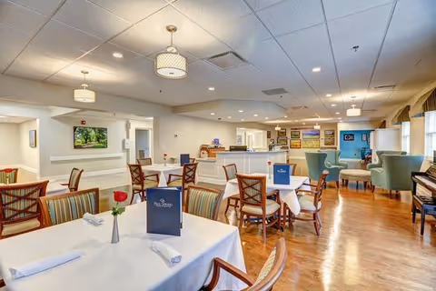 A bright and spacious dining room in a retirement community with several tables covered in white tablecloths, each set with napkins and a single red rose in a vase. The room features wooden chairs with striped cushions, soft lighting from ceiling fixtures, and a piano in the corner. Large windows with curtains allow natural light to fill the space.