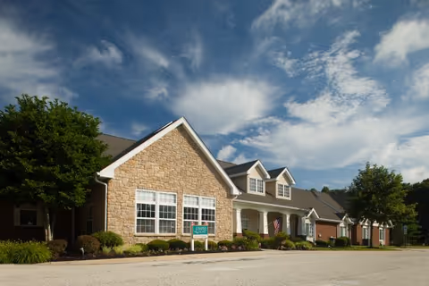 Front exterior of a single-story brick-and-stone senior living building with shrubs and an American flag beneath a blue sky.