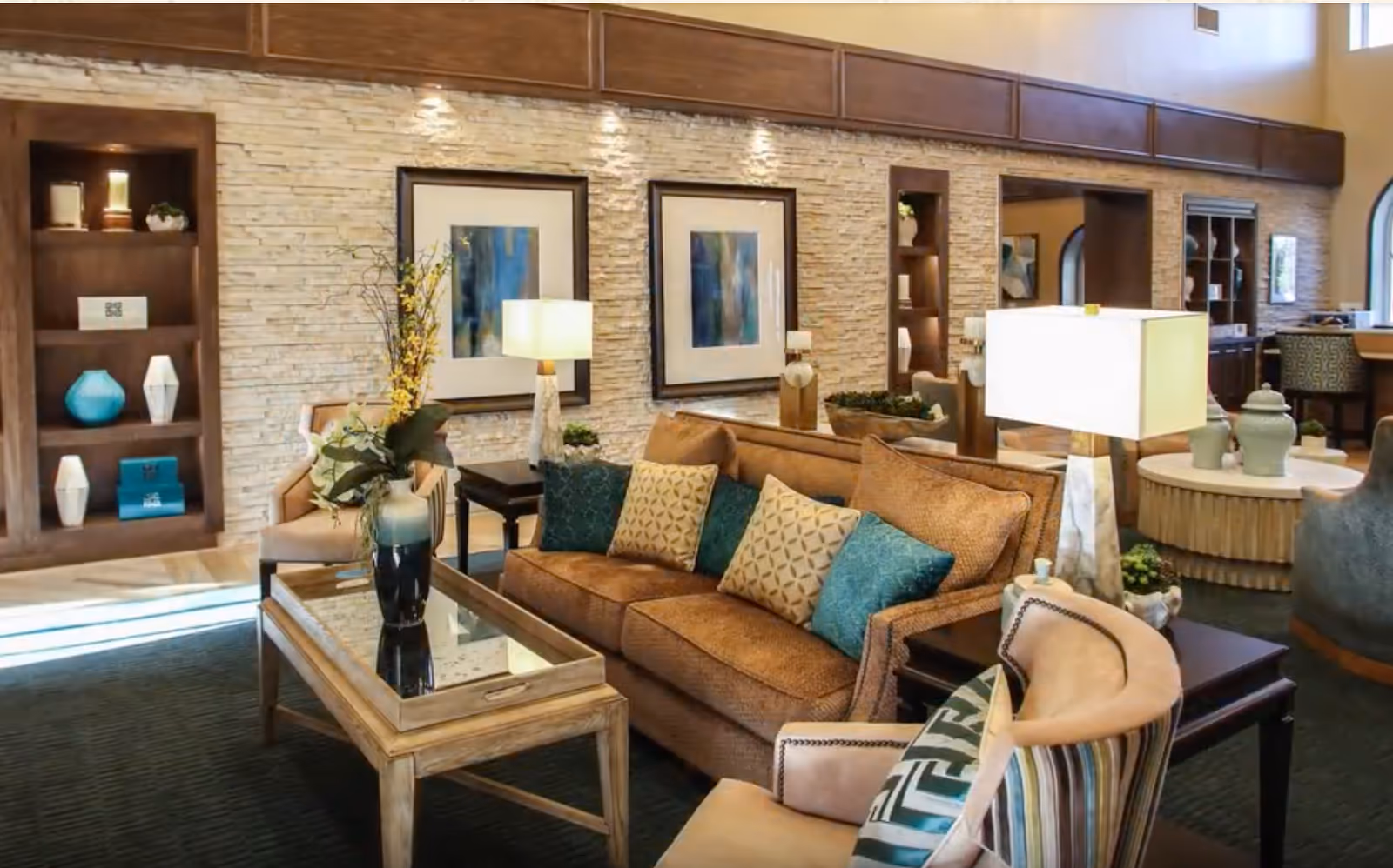 A bright, well-appointed communal living room with a sofa, armchairs, coffee table, lamps, and decorative shelving and artwork.