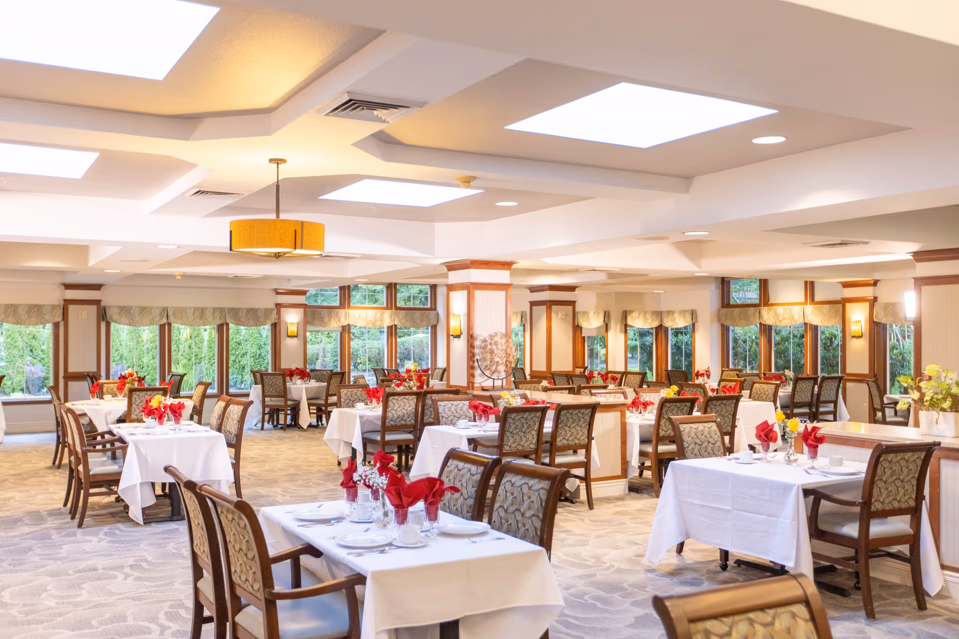 A bright and spacious dining room with multiple tables covered in white tablecloths, each set with plates, cups, and red napkins folded elegantly. The room features large windows with valances, allowing natural light to fill the space, and decorative ceiling lights. Chairs with patterned upholstery surround the tables, and there are floral centerpieces on some tables.