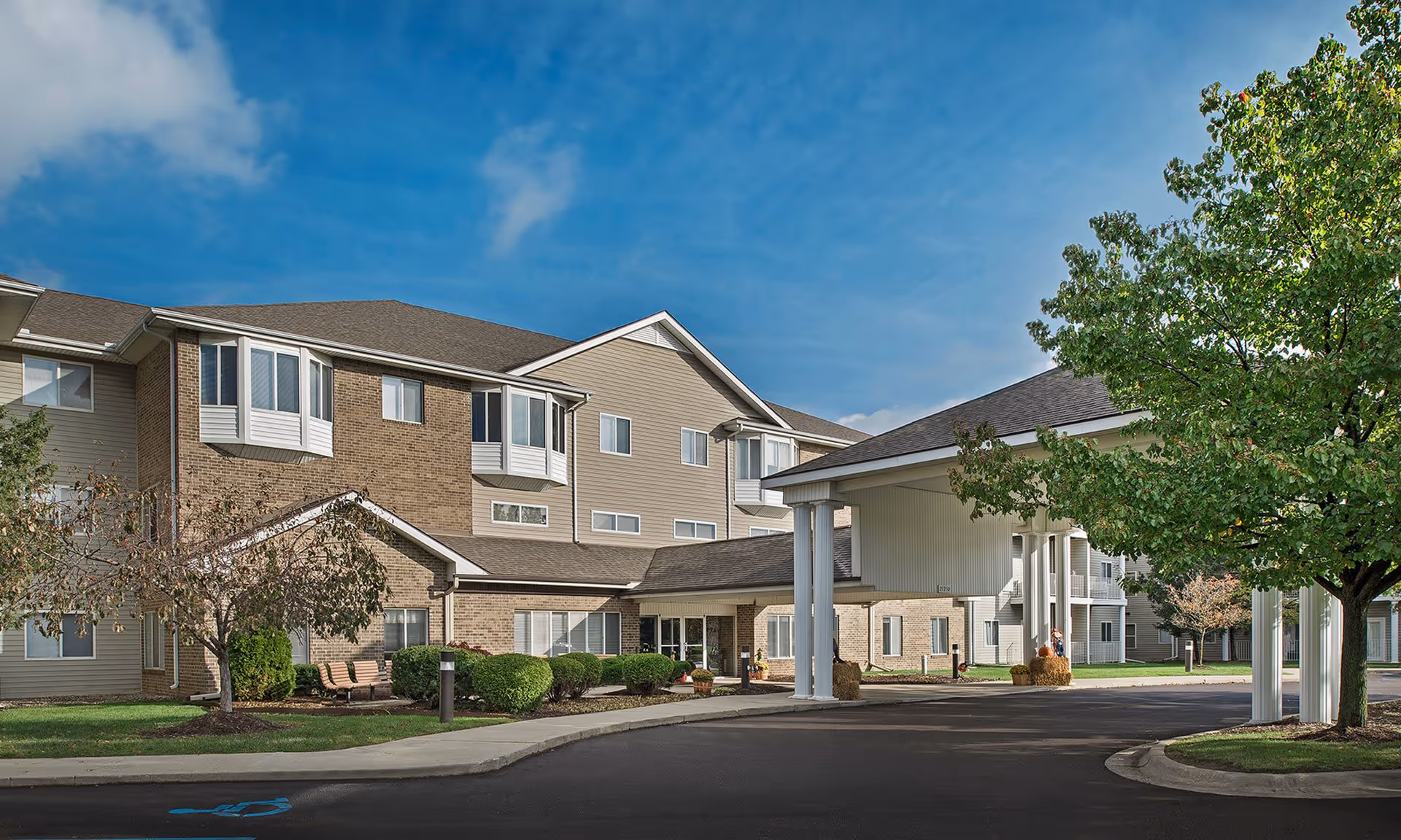 Exterior view of a senior living facility building with a covered entrance, surrounded by trees and landscaping under a blue sky.