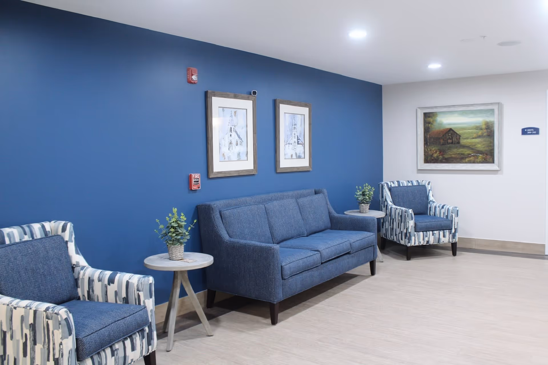 A seating area in a senior living facility with a blue accent wall, two framed pictures, a blue sofa, two patterned armchairs, two small round tables with potted plants, and a landscape painting on a white wall.