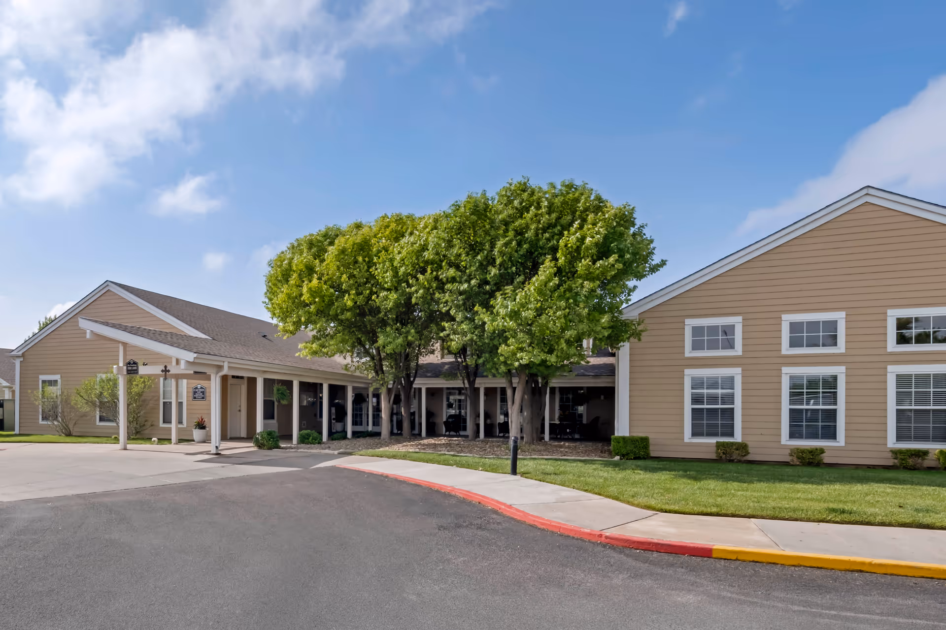 Exterior view of a single-story senior living facility building with beige siding, white trim, and multiple windows. There is a covered entrance with a small porch area, several green trees in front, a well-maintained lawn, and a paved driveway with a red and yellow curb under a partly cloudy blue sky.