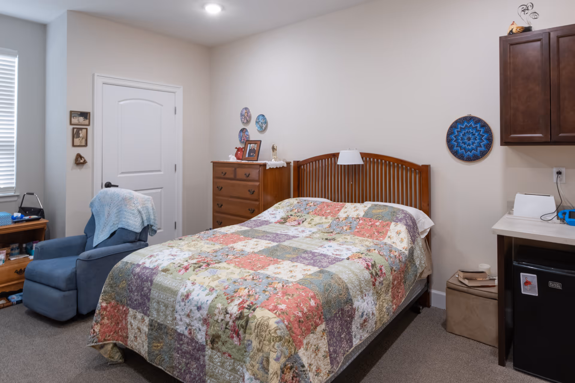 A bedroom with a wooden headboard and a colorful patchwork quilt on the bed, a blue recliner, dresser, and small cabinets with a mini fridge.