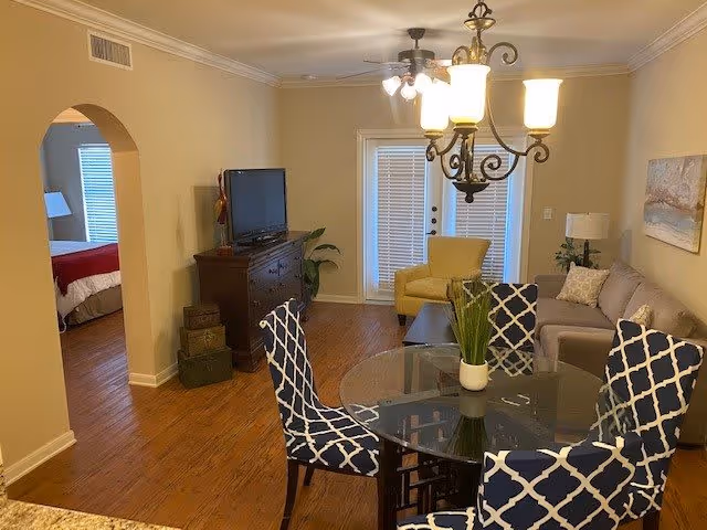 A cozy living room with a glass dining table surrounded by four blue and white patterned chairs. The room features a beige sofa with a pillow, a yellow armchair near glass doors with blinds, a dark wooden TV stand with a flat-screen TV, and a ceiling fan with lights. An archway leads to a bedroom with a red bedspread visible.