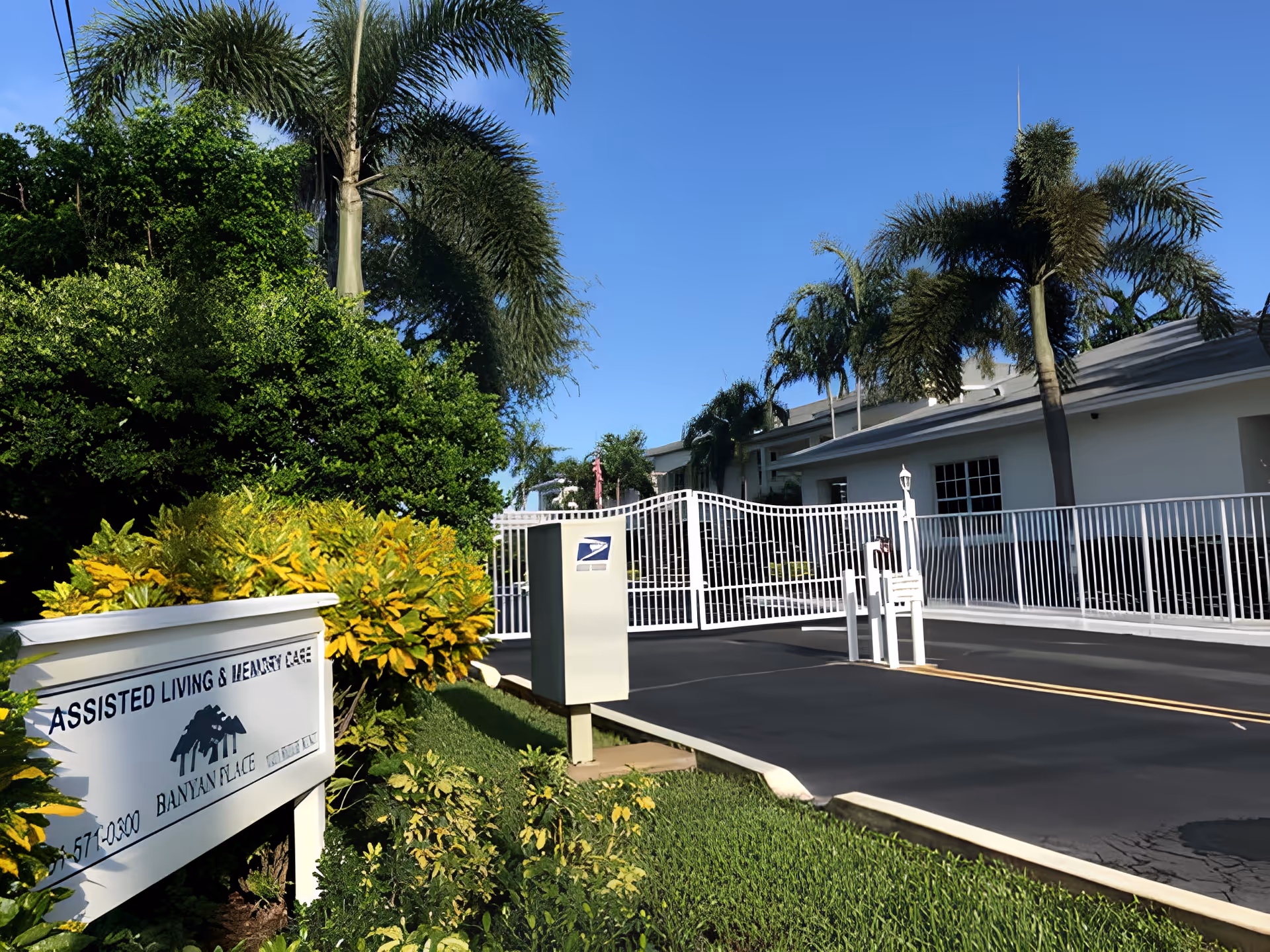Entrance to Banyan Place assisted living and memory care facility with a white gated driveway, lush green bushes, palm trees, and a clear blue sky.