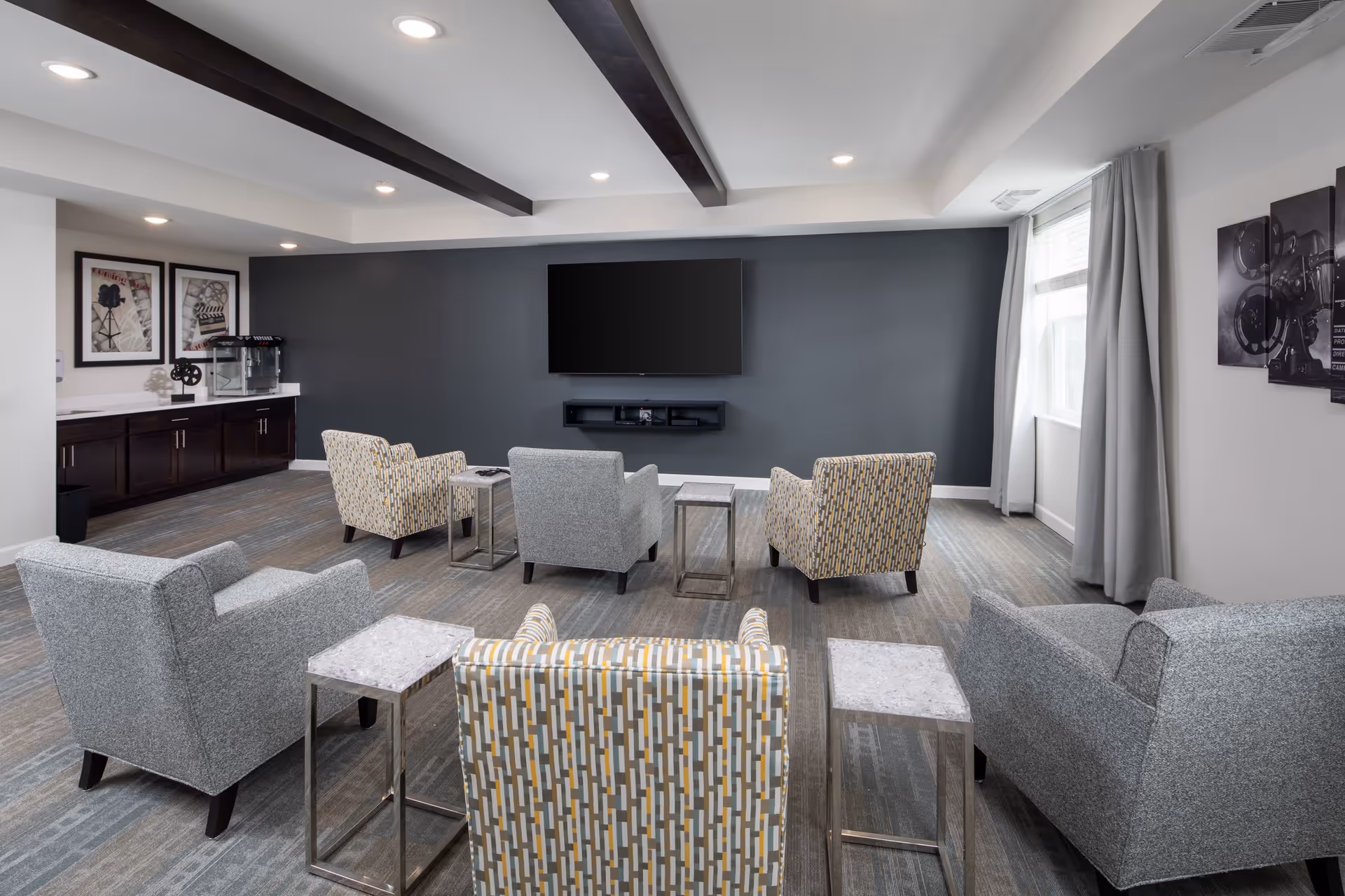 A modern living room with six armchairs arranged in two rows facing a large flat-screen TV mounted on a dark gray accent wall. The room has a light gray carpet, white walls, ceiling with recessed lighting, and dark wooden beams. There are small side tables between the chairs, framed artwork on the walls, and a window with gray curtains letting in natural light.